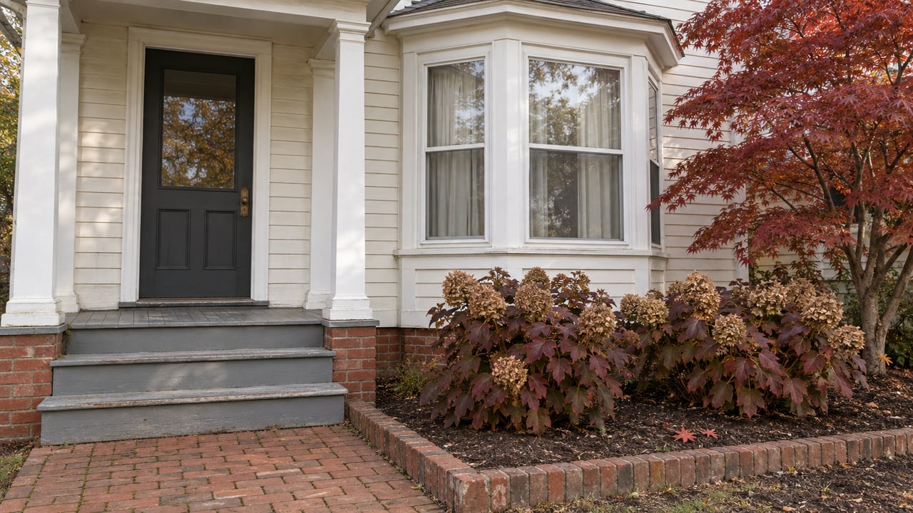 White clapboard home with a charcoal entry door, glazed bay window, brick walk and copper-red autumn planting.