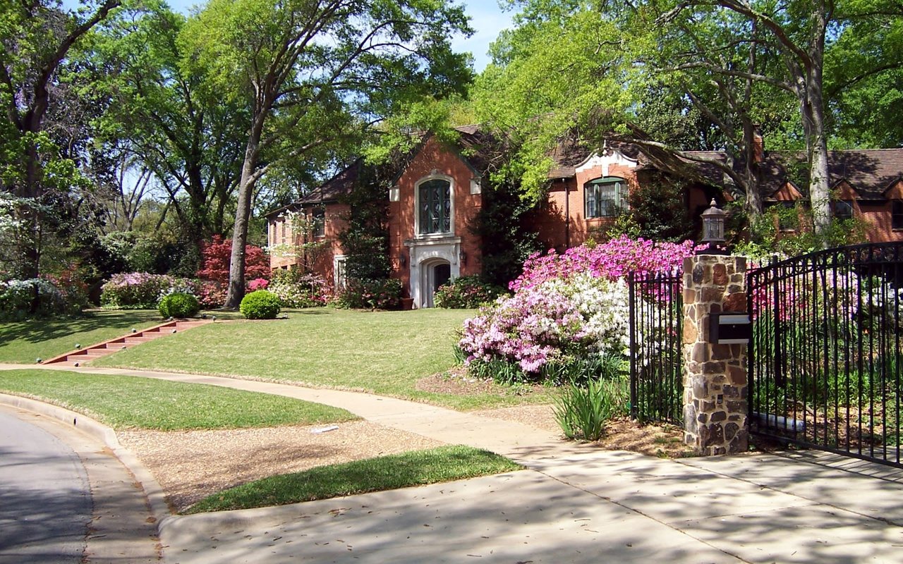 Red-brick house behind a lawn, flowering shrubs, mature trees, and a black metal gate.