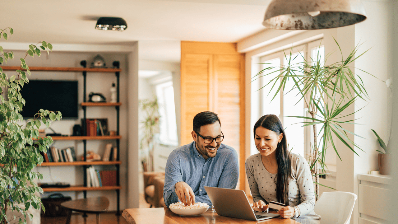 A smiling couple sits at a kitchen table in a bright, modern home. The man wears glasses and a blue shirt, while the woman in a light gray sweater types on a laptop, searching for homes for sale in Gainesville FL. Gainesville FL realtor Anna Olcese.