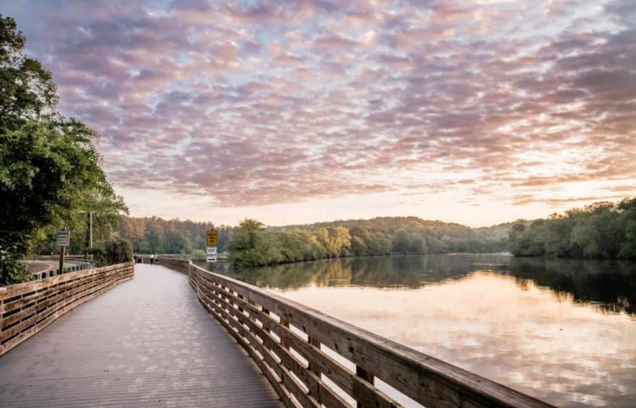 Wooden boardwalk beside a calm river beneath a pink and gold sky.