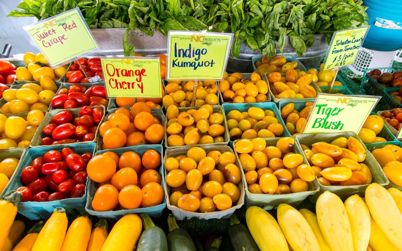 Farmers market display of colorful tomatoes and squash with handwritten variety signs.