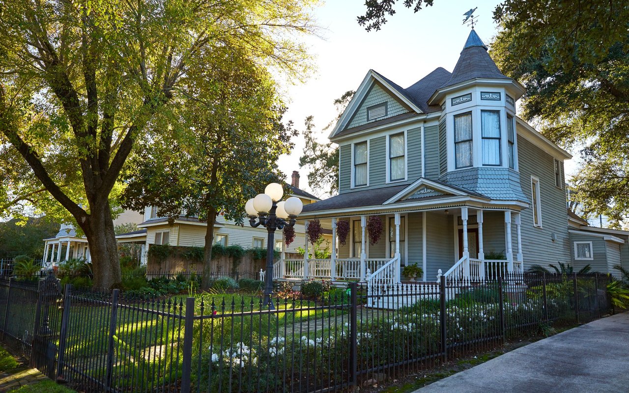 Blue-gray Victorian house with a turret, wraparound porch, and fenced landscaped yard.