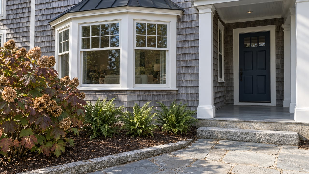 Weathered cedar-shingled home with white-trimmed bay windows, a sheltered navy entry door, granite steps and burgundy oakleaf hydrangea.