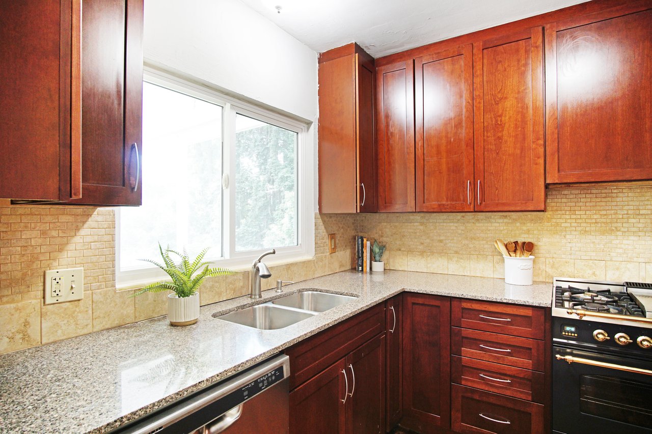 Warm kitchen with cherry wood cabinets and a grey granite countertop. Features a double sink, gas stove, and a potted fern by a sunny window.