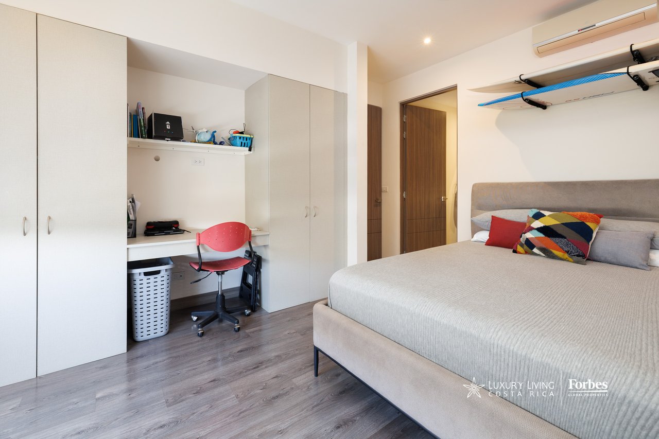Bedroom with gray bed, built-in wardrobes, desk, red chair, and surfboards stored overhead.