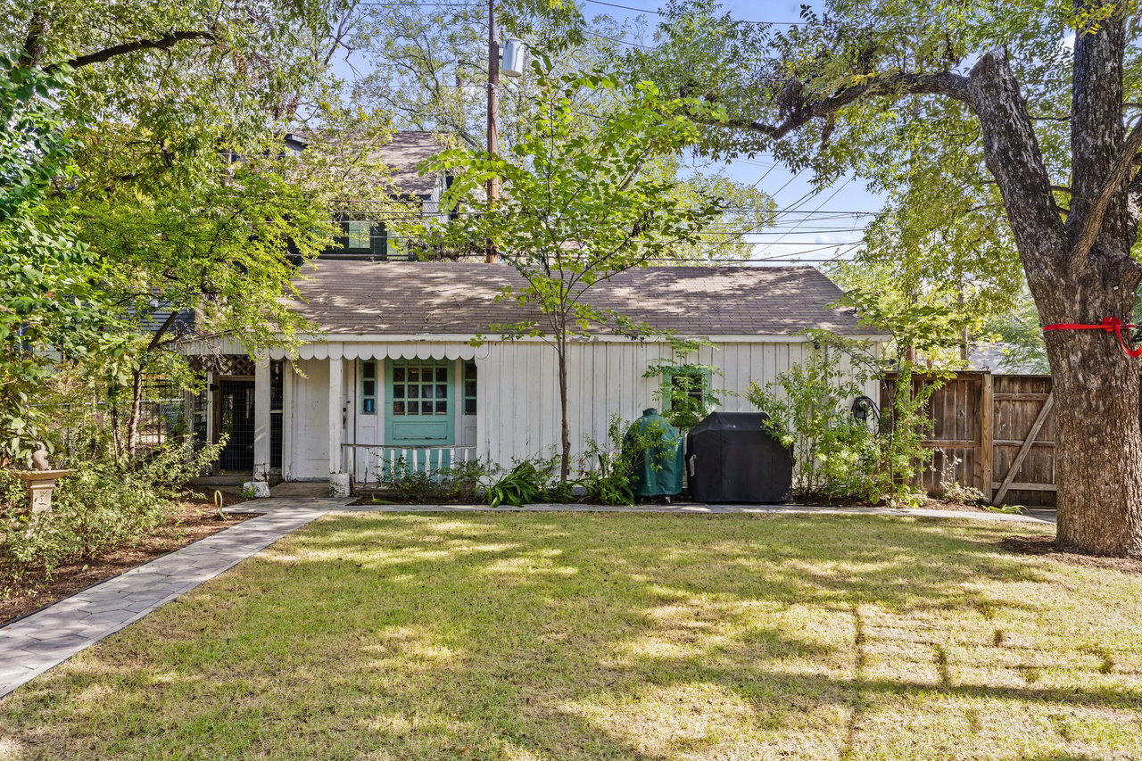 White home's rear exterior with turquoise door, scalloped awning, and a tree-shaded lawn.