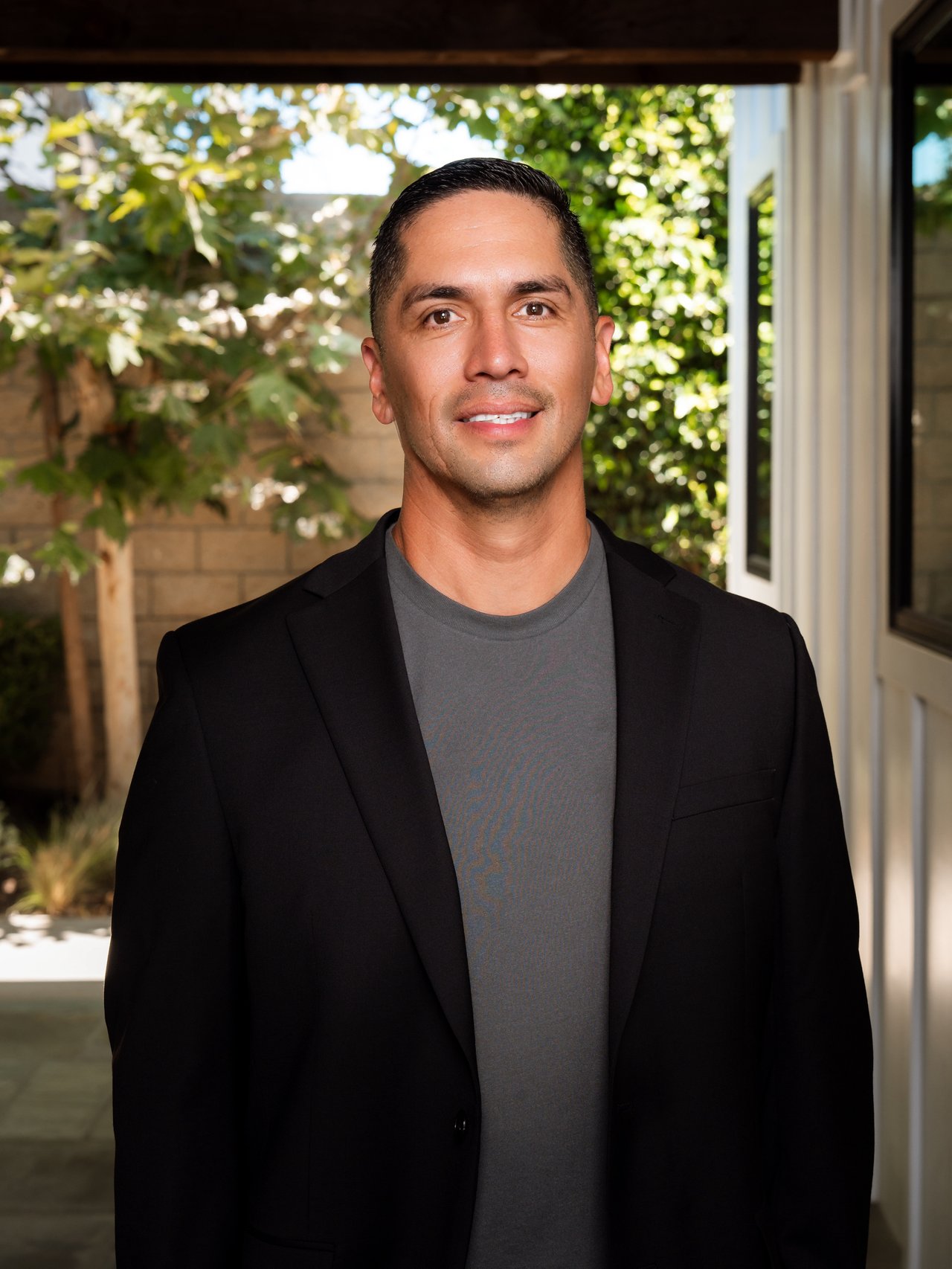 Brian Zambrano in a black blazer and gray shirt, standing in front of leafy greenery.