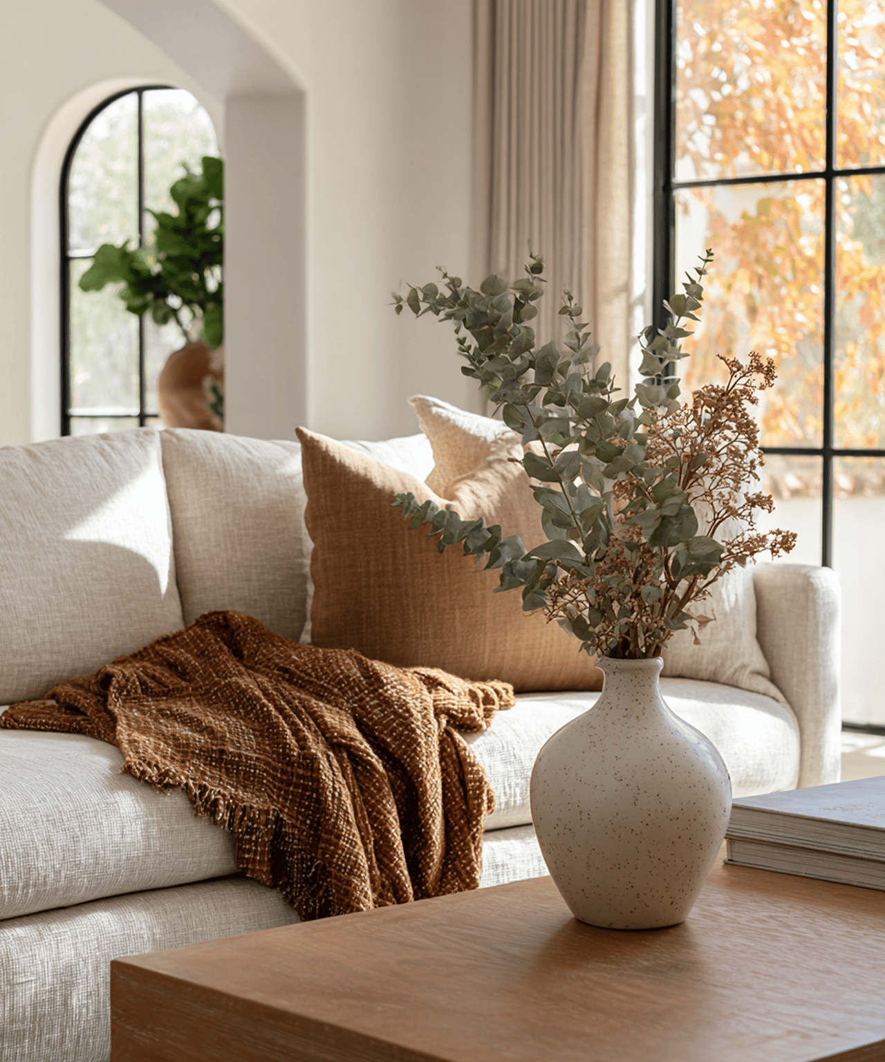 Staged living room with a cream sofa, brown throw, and vase of branches by a sunlit window.