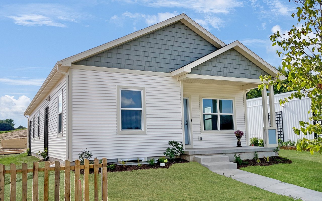 White single-story house with a covered porch, gray gabled roof, and wooden picket fence.
