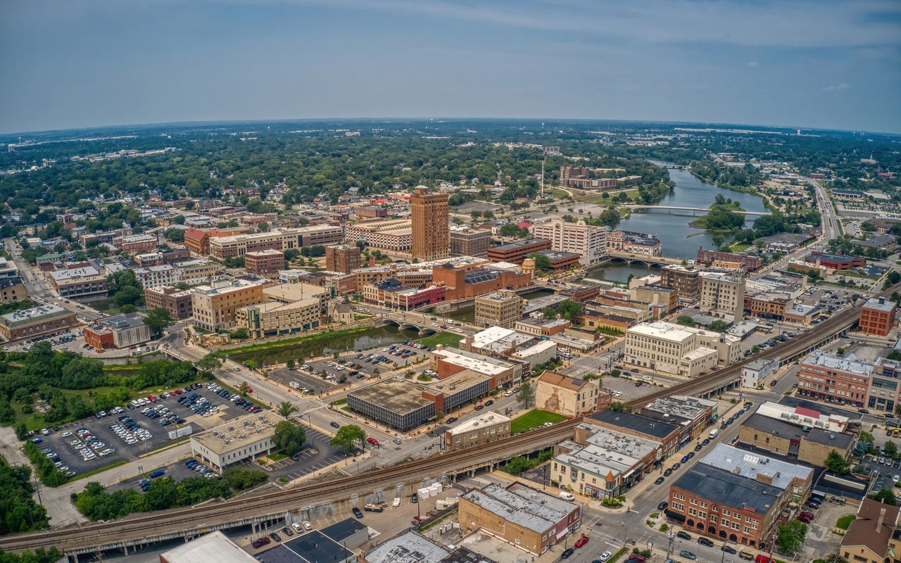 Aerial panorama of a downtown beside a river, with bridges, railway tracks, and green neighborhoods.
