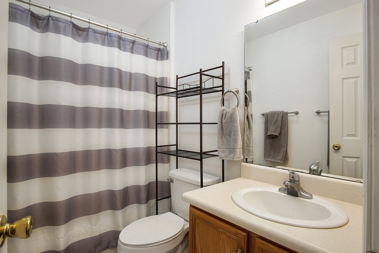 A small bathroom with a beige countertop and sink, striped shower curtain, white toilet, metal shelf, and gray towels, creating a clean and minimal look.