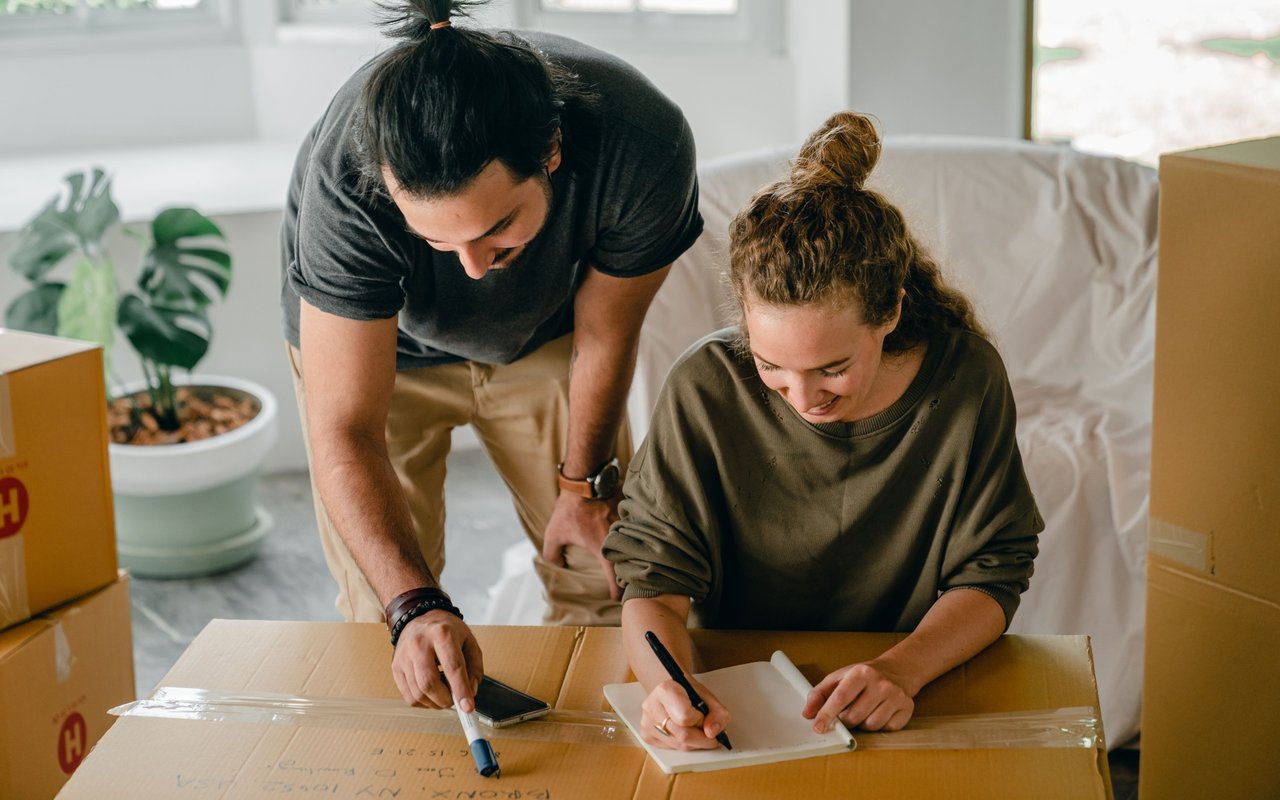 A couple writes on a moving box while packing their belongings in a bright living room.