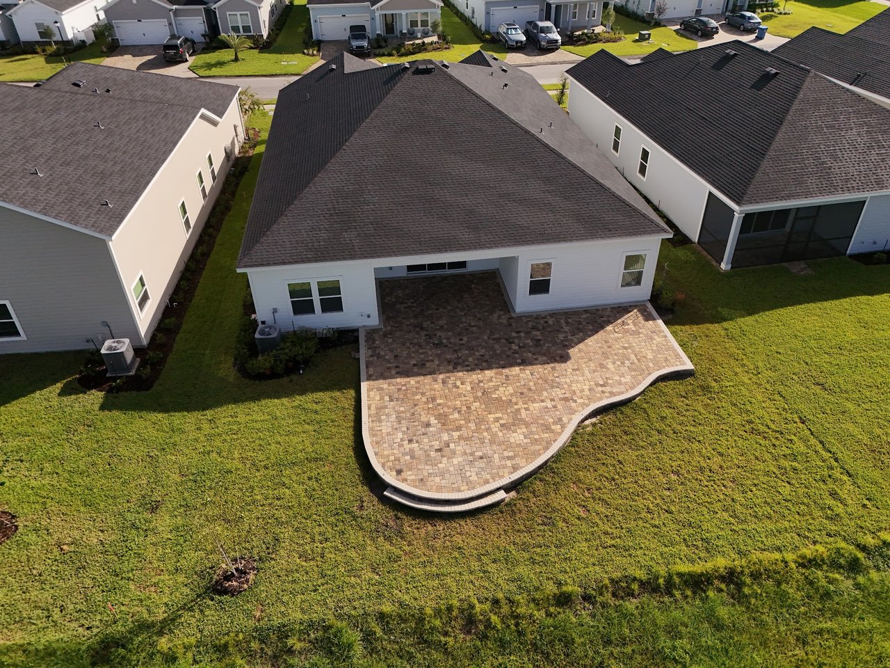 Aerial view of a white home’s backyard, with a curved paver patio and green lawn.