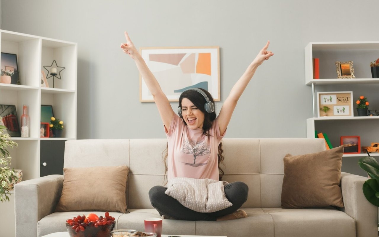 Woman wearing headphones celebrates with her arms raised while sitting on a living room sofa.