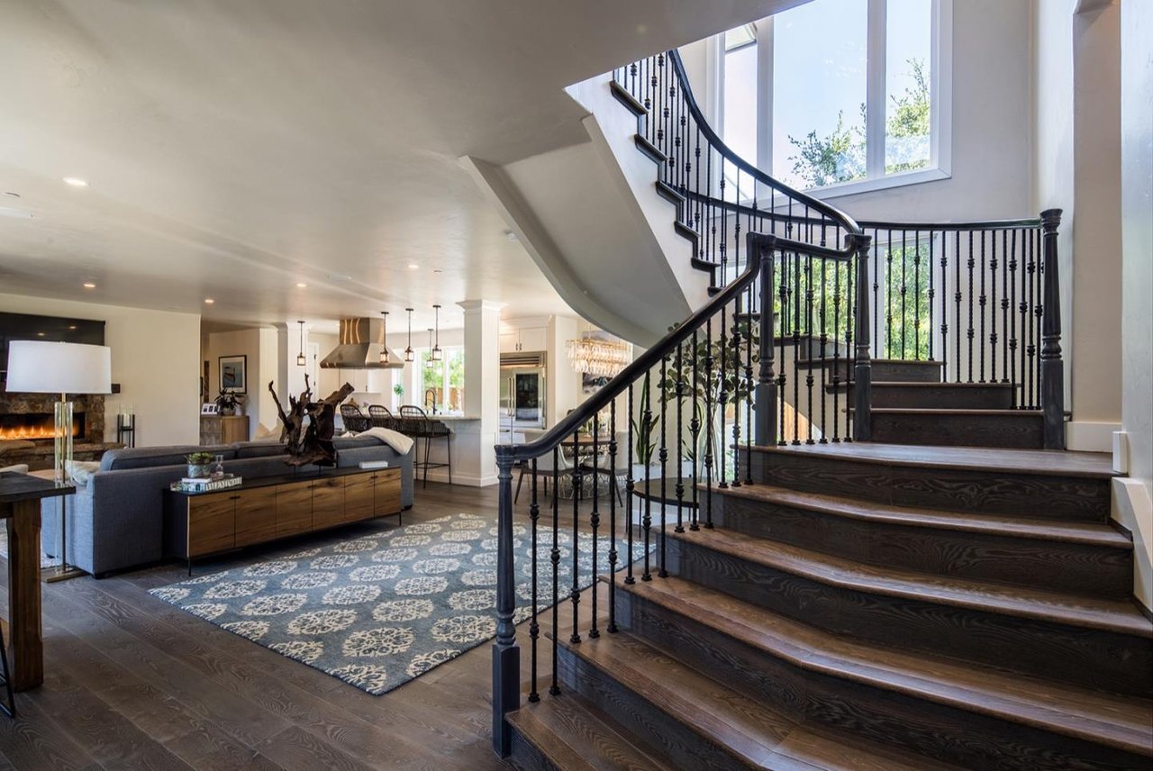 Open-plan living room with a sweeping dark wood staircase, black iron railing, and stone fireplace.