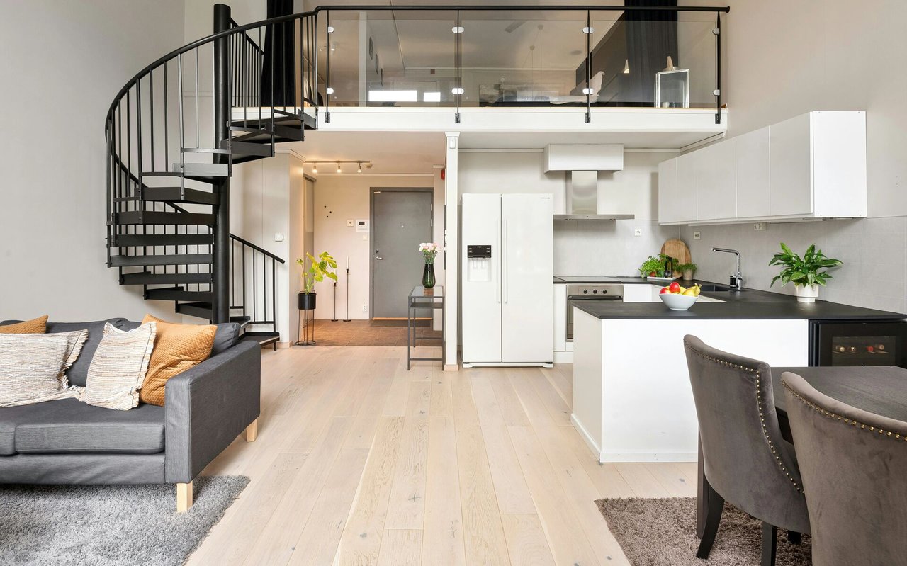 Open-plan loft with a black spiral staircase, glass-railed mezzanine, gray sofa, and white kitchen.
