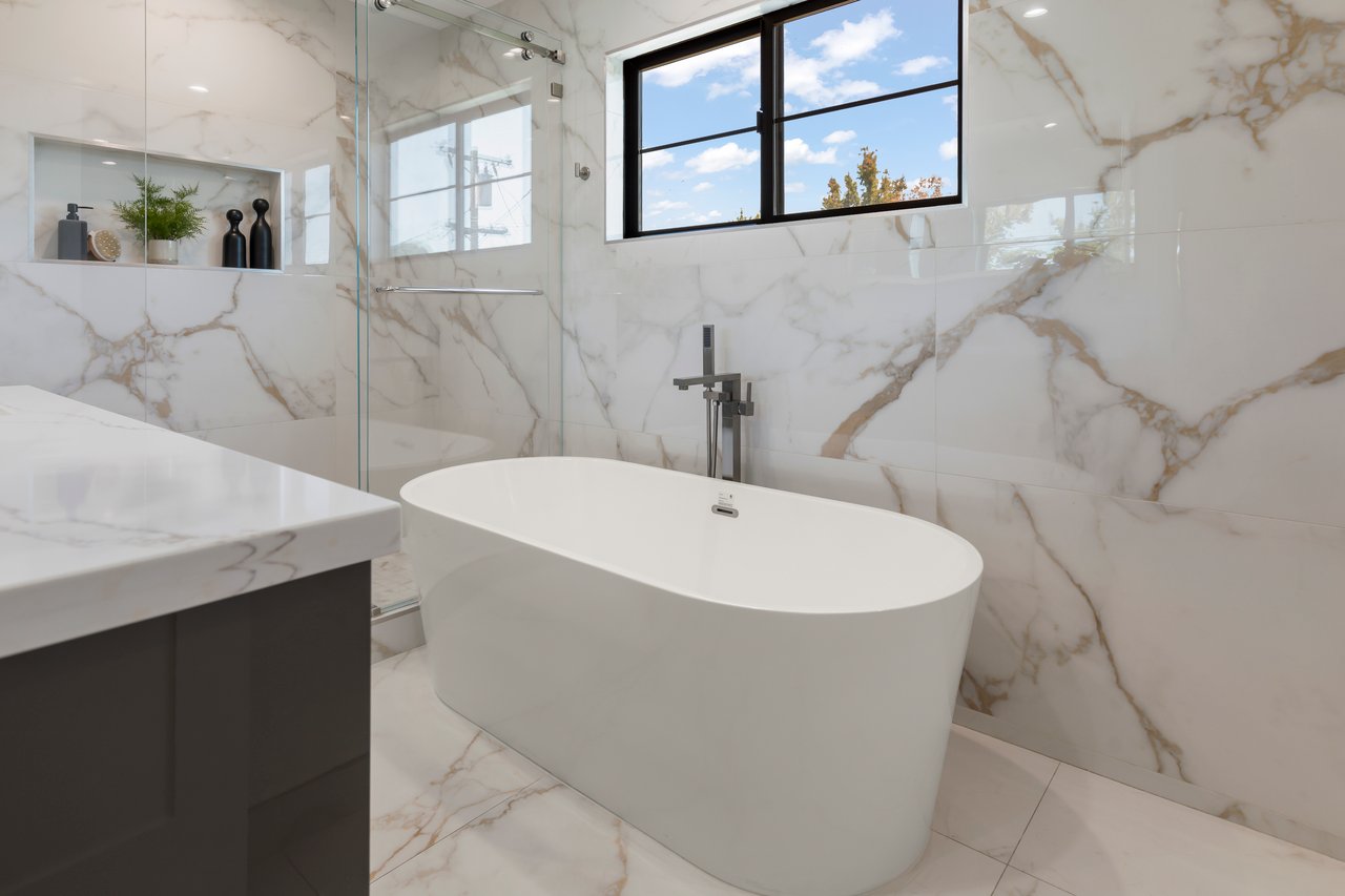 Bathroom with a freestanding soaking tub, marble-look walls, and a black-framed window.