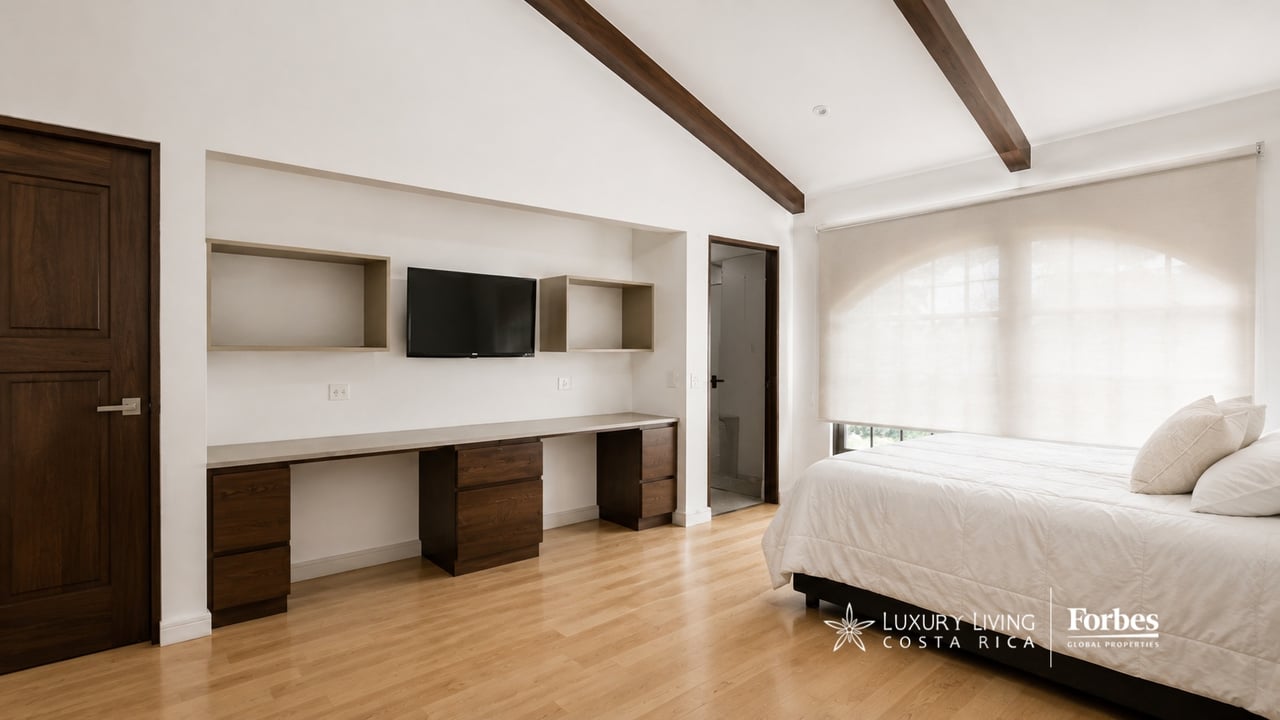 Bedroom with a white bed, wood floors, ceiling beams, and a built-in desk and television.