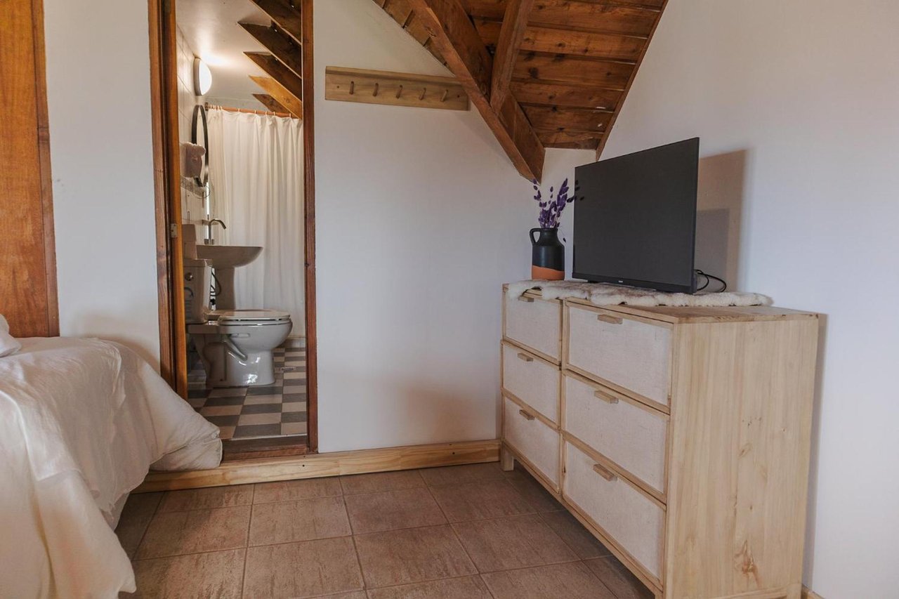 Hotel bedroom with exposed timber ceiling, wooden dresser and an adjoining bathroom visible through an open doorway.
