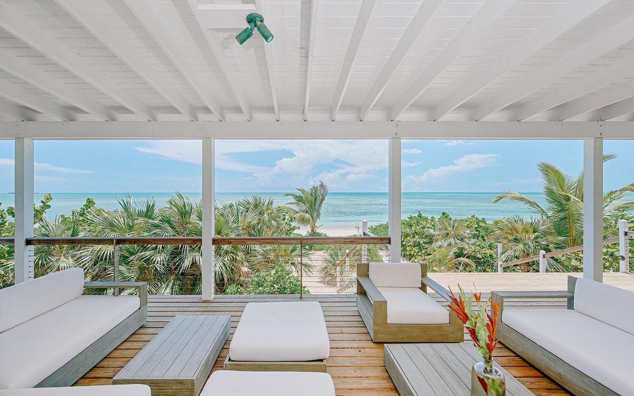 Covered oceanfront patio with white lounge seating, a wood deck, and palm trees.