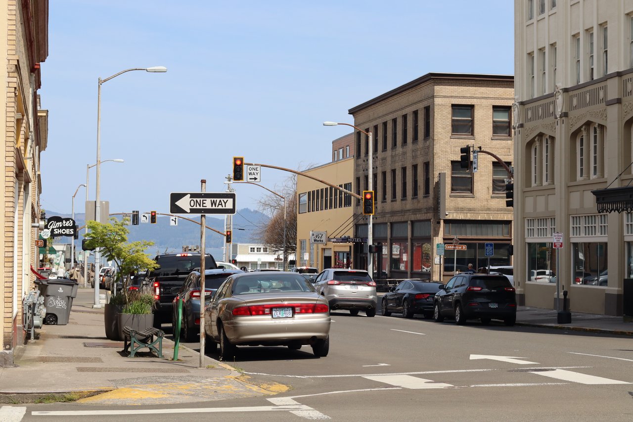 Downtown Astoria street intersection with historic commercial buildings, parked and moving cars, traffic lights, and one-way signs under a clear blue sky. Mountains are visible in the distance.