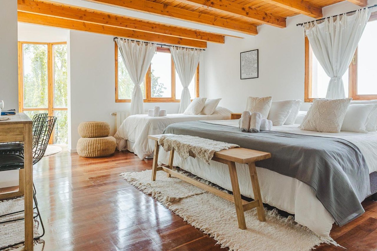Hotel bedroom with two beds, exposed timber ceiling beams, and large curtained windows.