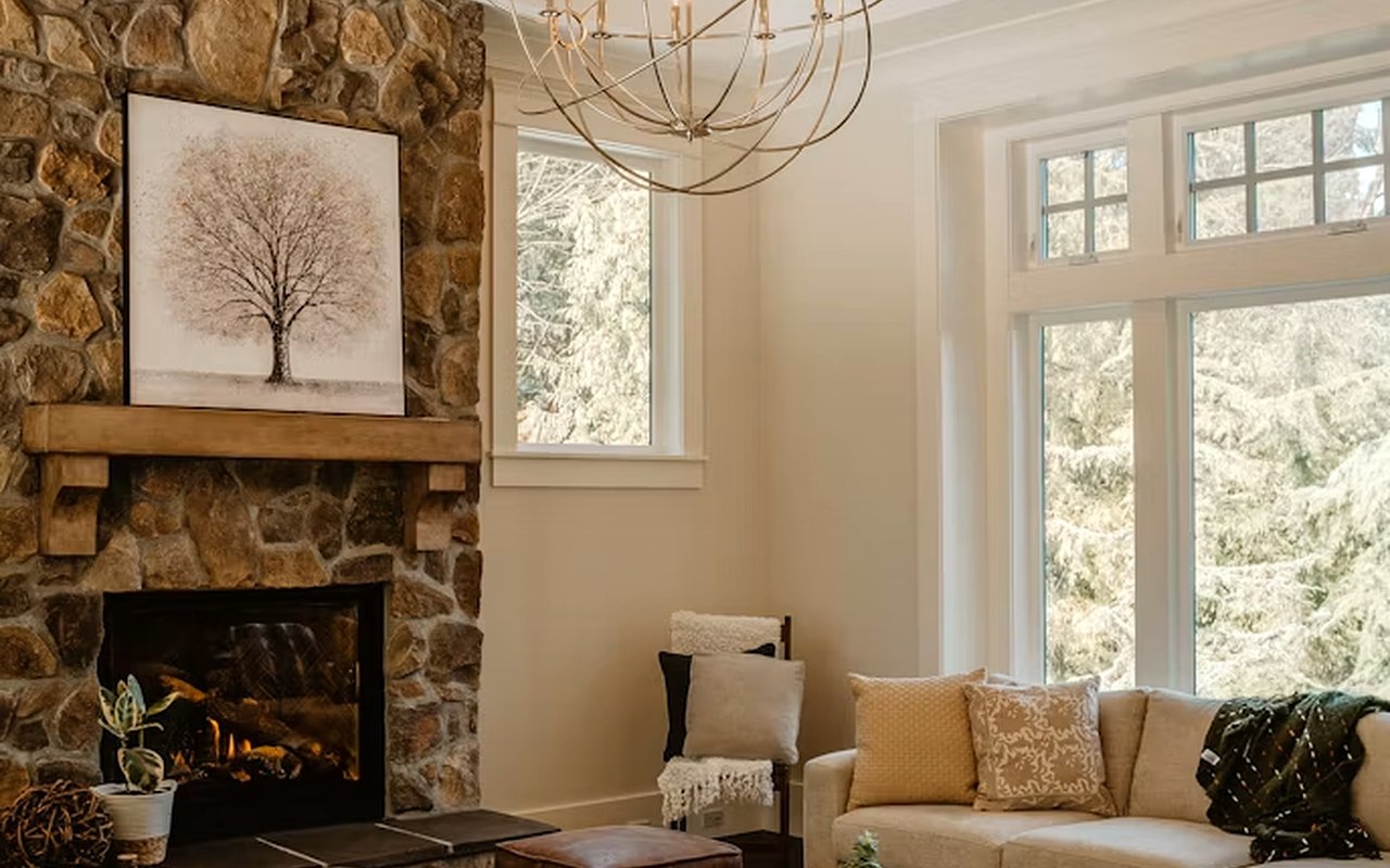 Living room with a stone fireplace, beige sofa, and large windows overlooking snowy evergreens.