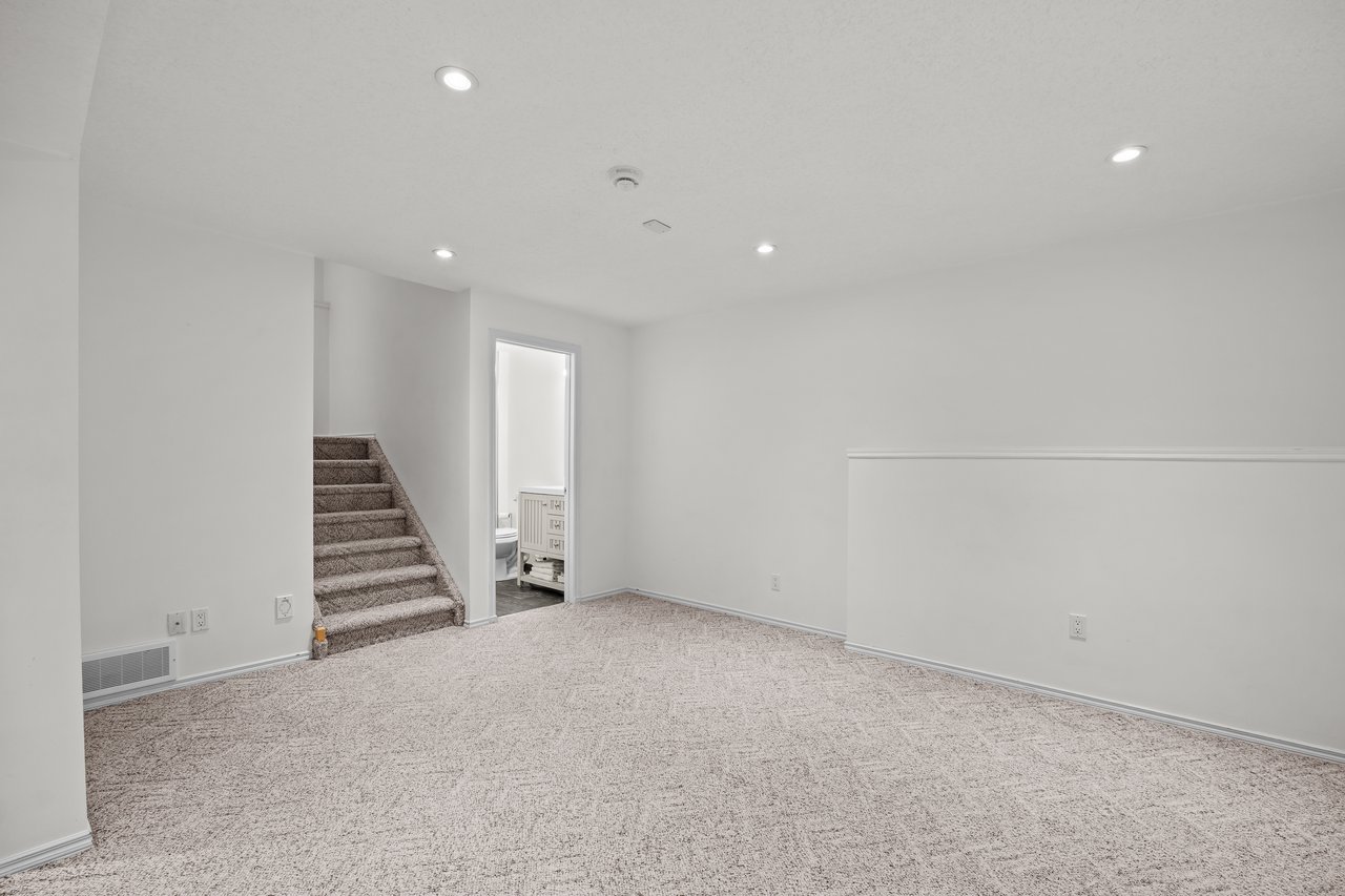 Empty carpeted basement room with recessed lights, stairs, and an open bathroom doorway.