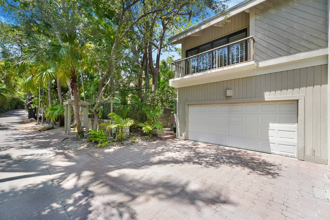 Two-story gray home with a double garage, upper balcony, and tropical landscaping.