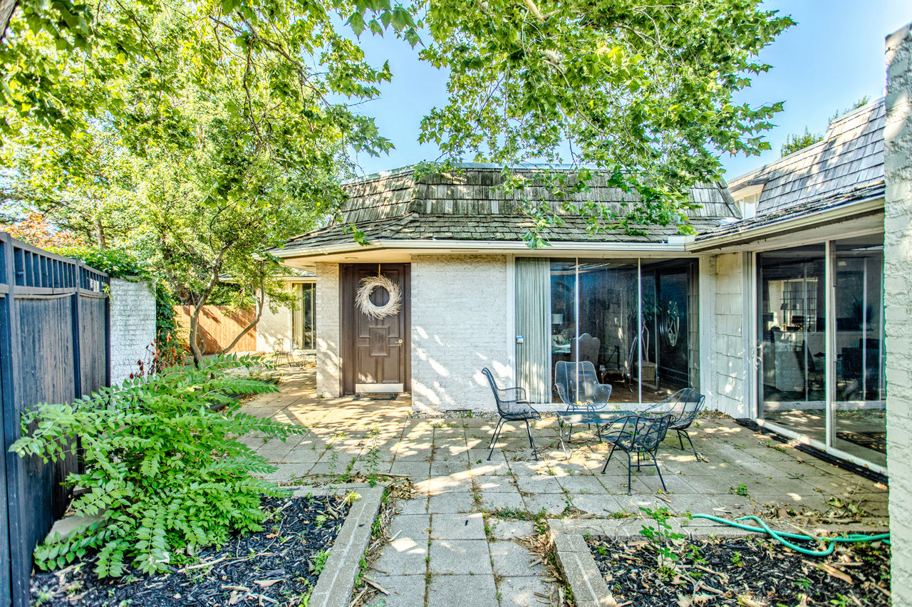 Home’s shaded rear patio with sliding glass doors, outdoor seating, and mature trees.