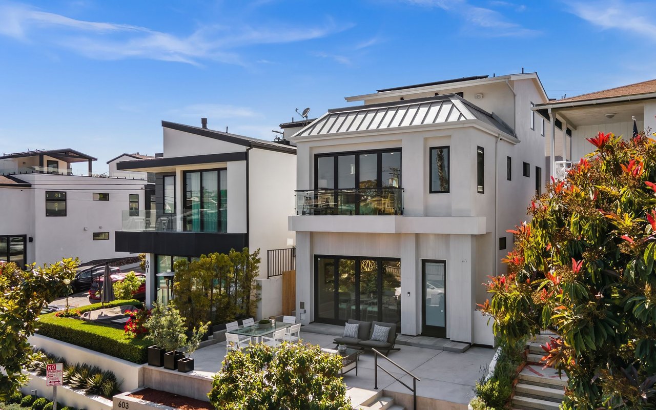 Modern white home with a metal roof, glass balcony, and furnished patio among landscaped gardens.