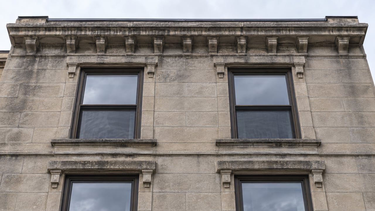 Weathered limestone condominium facade with four dark-framed sash windows and a low roof parapet beneath pale sky.