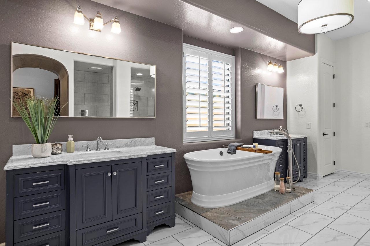 Spacious primary bathroom with dark dual-sink vanity, large mirrors, freestanding soaking tub on a tiled platform, and window shutters.