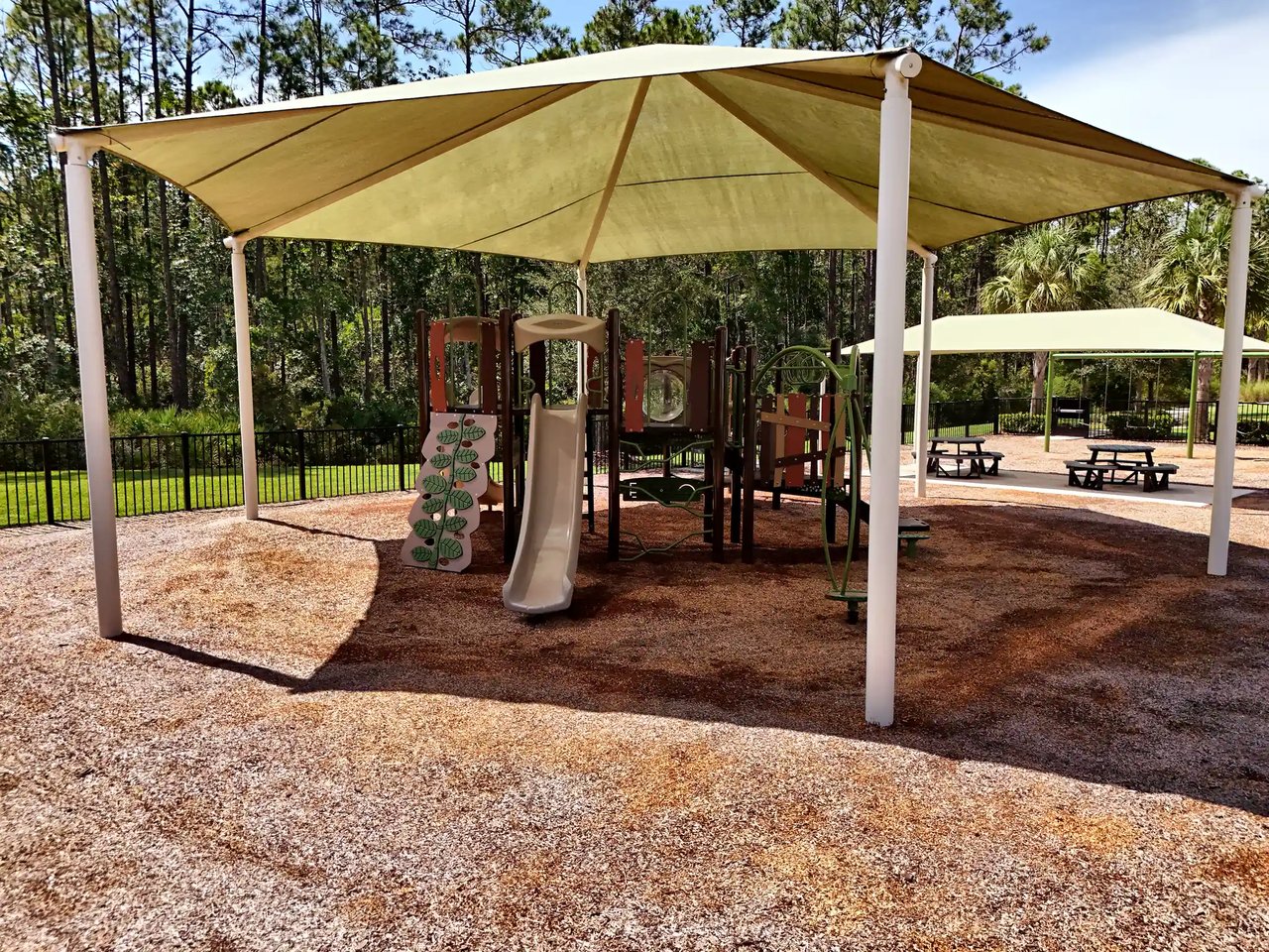 Playground with slides and climbing equipment under a large shade canopy, with picnic tables and pines behind.