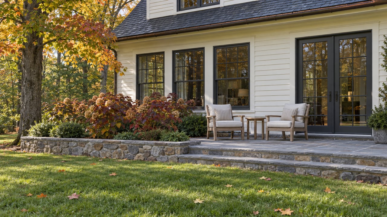 An autumn lawn leads to a bluestone terrace with two teak chairs beside a cream clapboard home and a maple turning ochre.