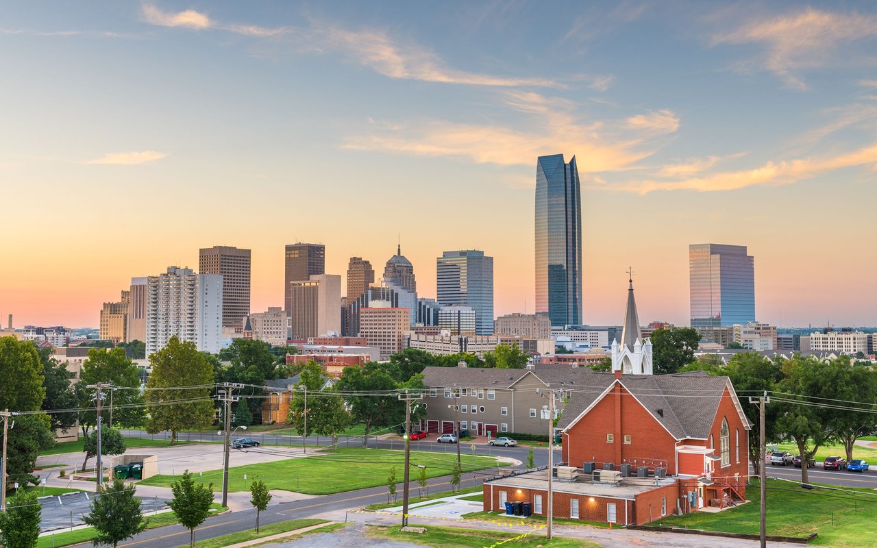 Oklahoma City skyline at sunset, with downtown towers and a red-brick church in the foreground.