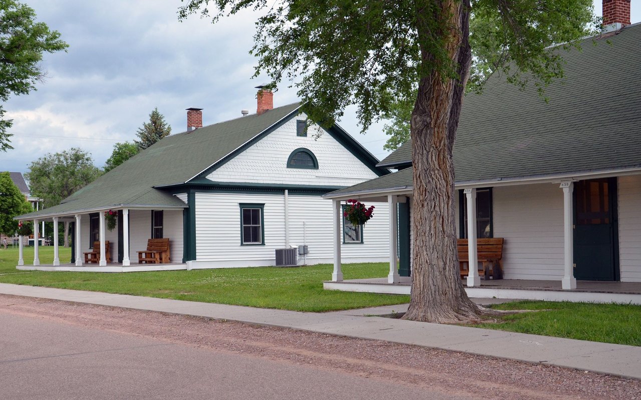 White buildings with green trim and covered porches beside a large tree.