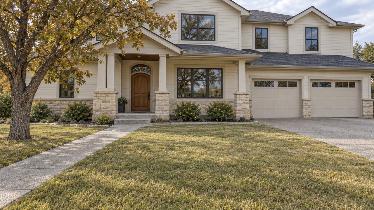 A two-story cream-sided home with a recessed oak entry, stone porch, attached garage and deep lawn beneath an autumn oak.