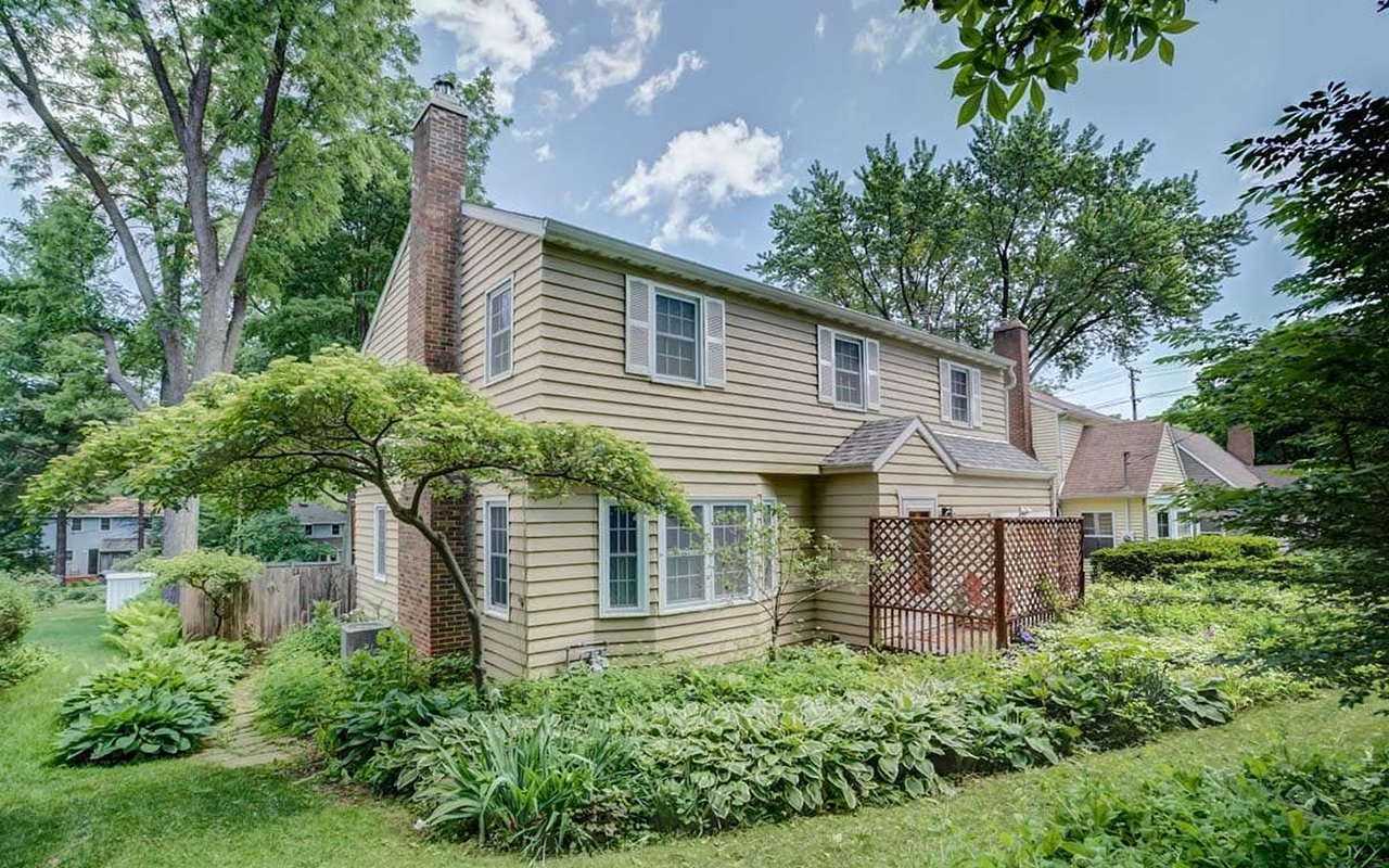 Two-story beige house with brick chimneys, a lattice screen, and mature trees and garden beds.