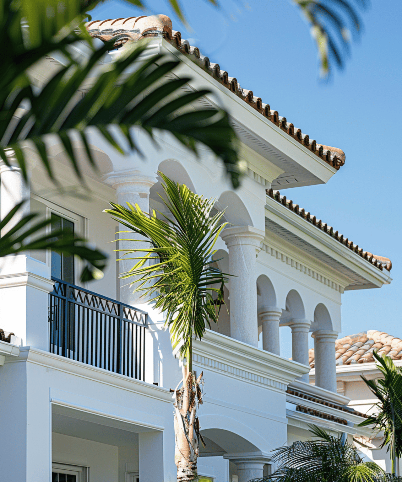 White Mediterranean-style home with arched balconies, columns, terracotta roof tiles, and palm trees.