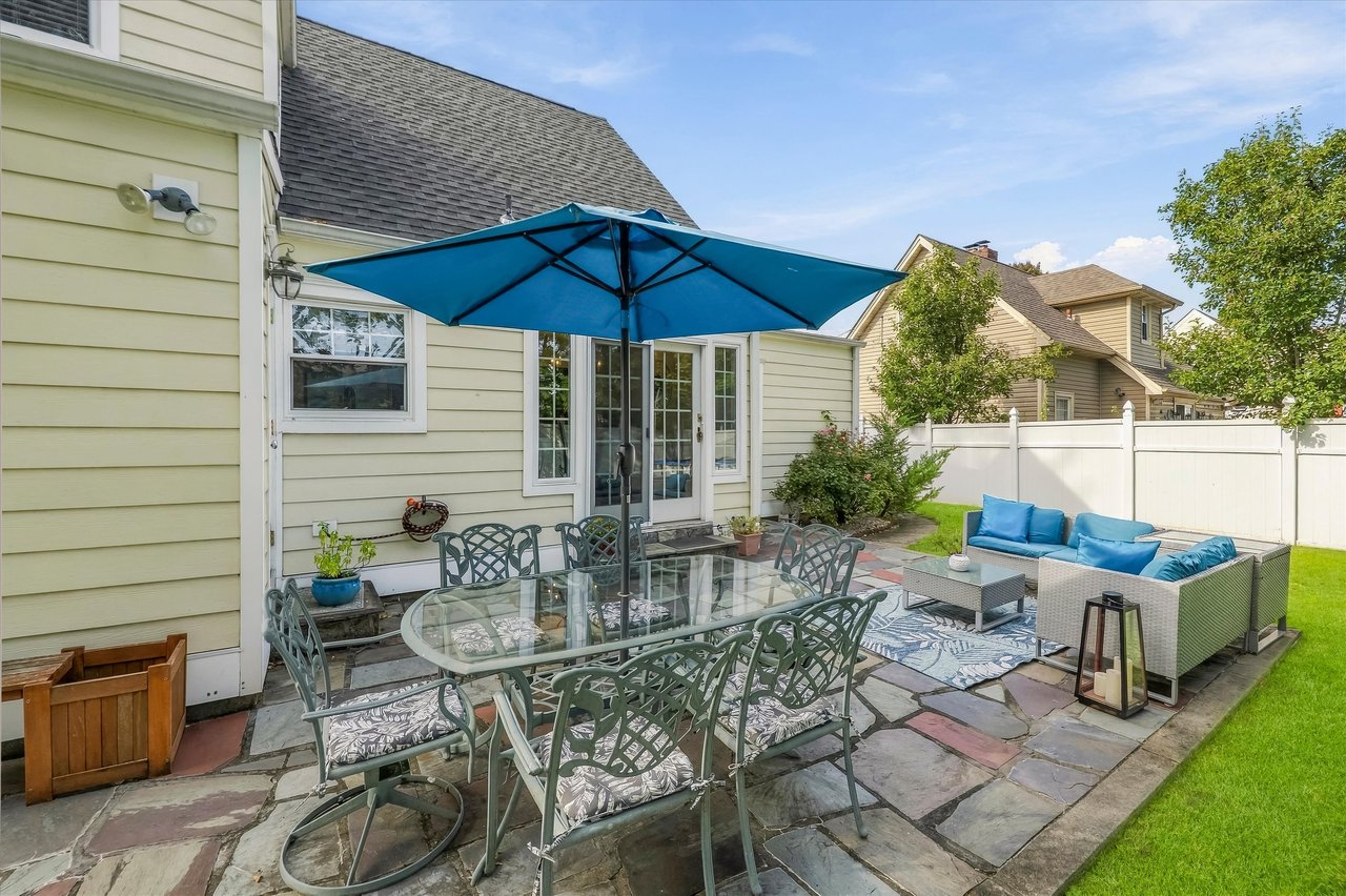 Backyard patio with a blue umbrella over a glass dining table and cushioned seating beside the house.