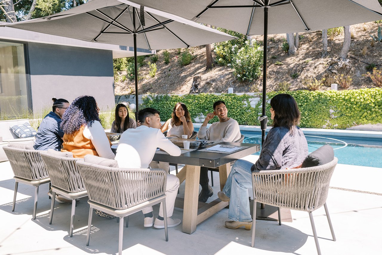 Seven adults gather around a patio table beside a swimming pool.