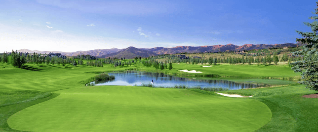 Golf course with a foreground putting green, pond, sand bunkers, and mountains in the distance.