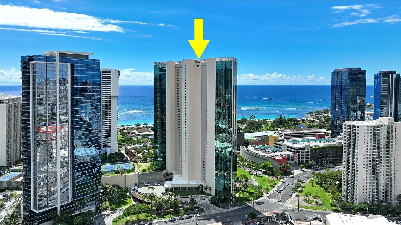 High-rise condominium tower marked by an arrow, with ocean and neighboring buildings behind it.