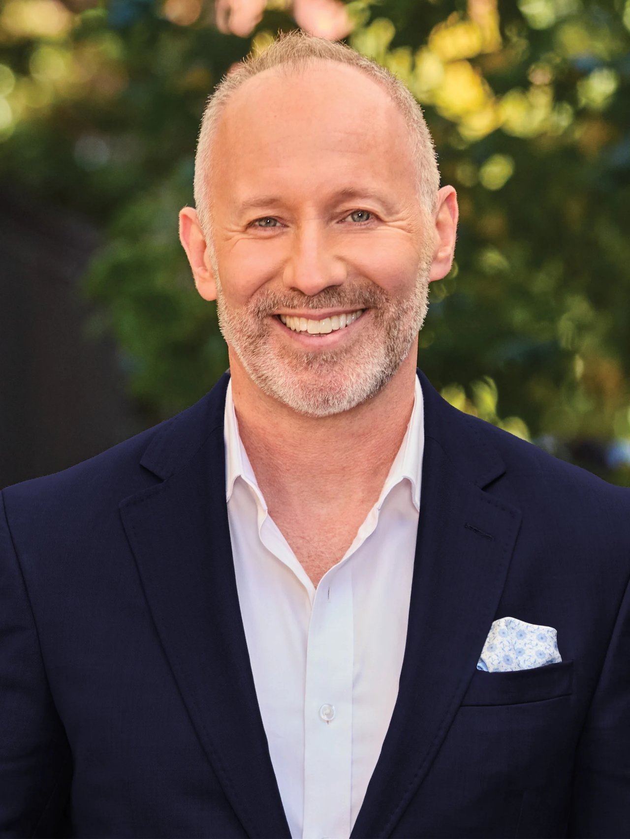 Steven Cohen smiling in a navy suit and white shirt against blurred greenery.