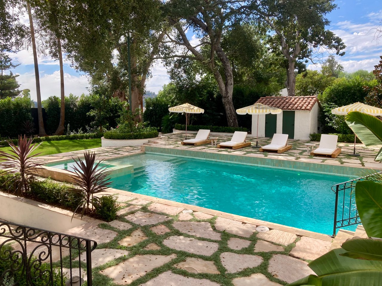 Lush Spanish-inspired LA backyard with sparkling pool, flagstone deck, yellow-striped umbrellas, and white stucco cabana.