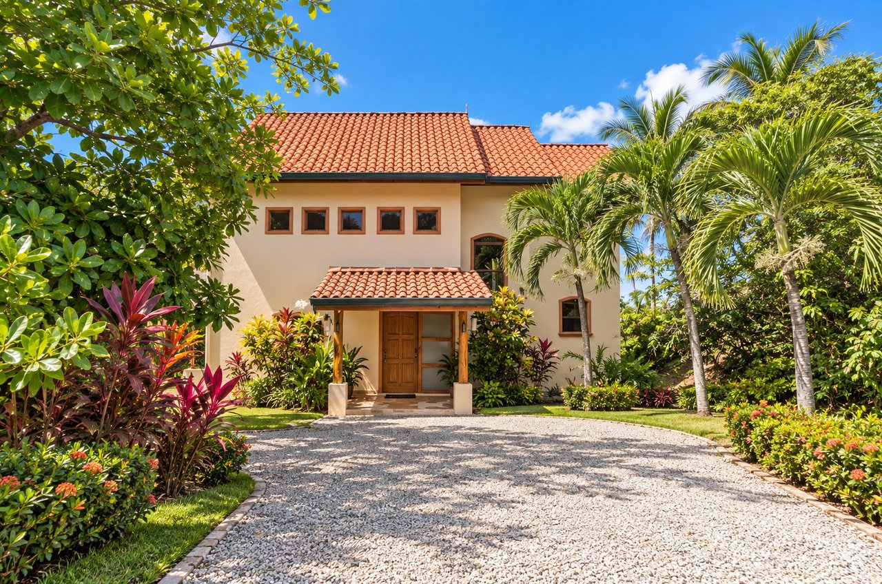 Two-story cream house with terracotta roofs, tropical landscaping, and a curved gravel driveway.