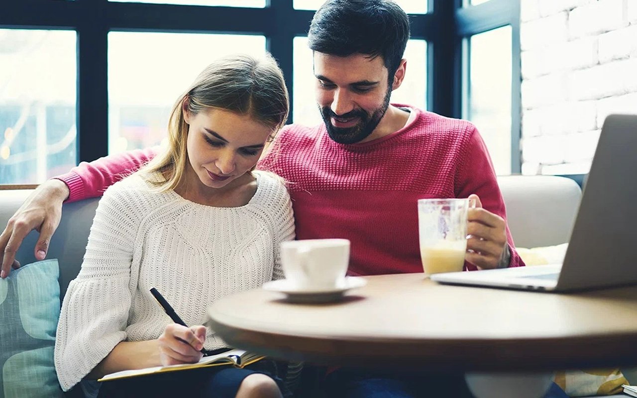 A couple reviews a notebook together at a café table beside coffee and a laptop.