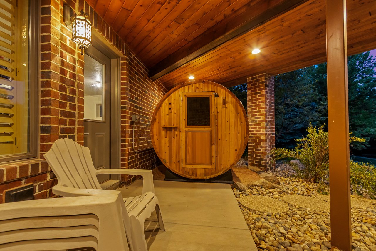 Covered brick patio with a round wooden barrel sauna and white Adirondack chairs.