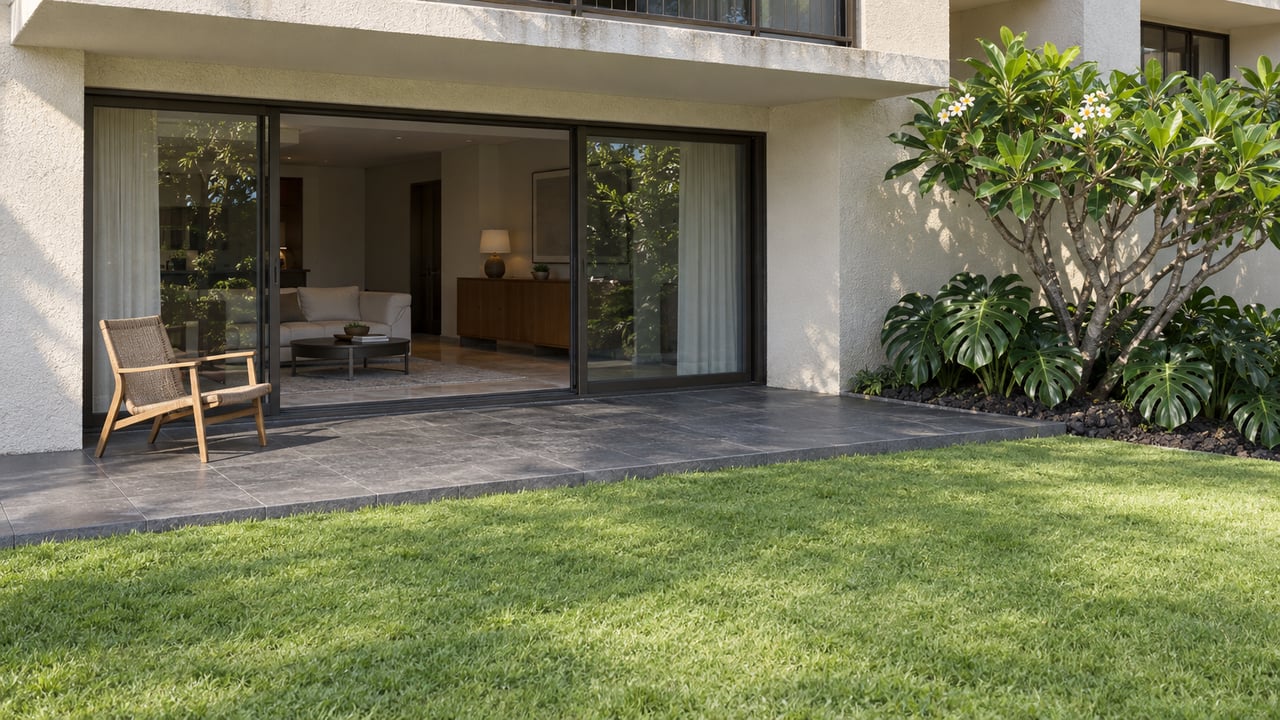 Elevated view of a stone-paved condominium terrace with a teak chair, enclosed sliding glass doors, plumeria and a green lawn.